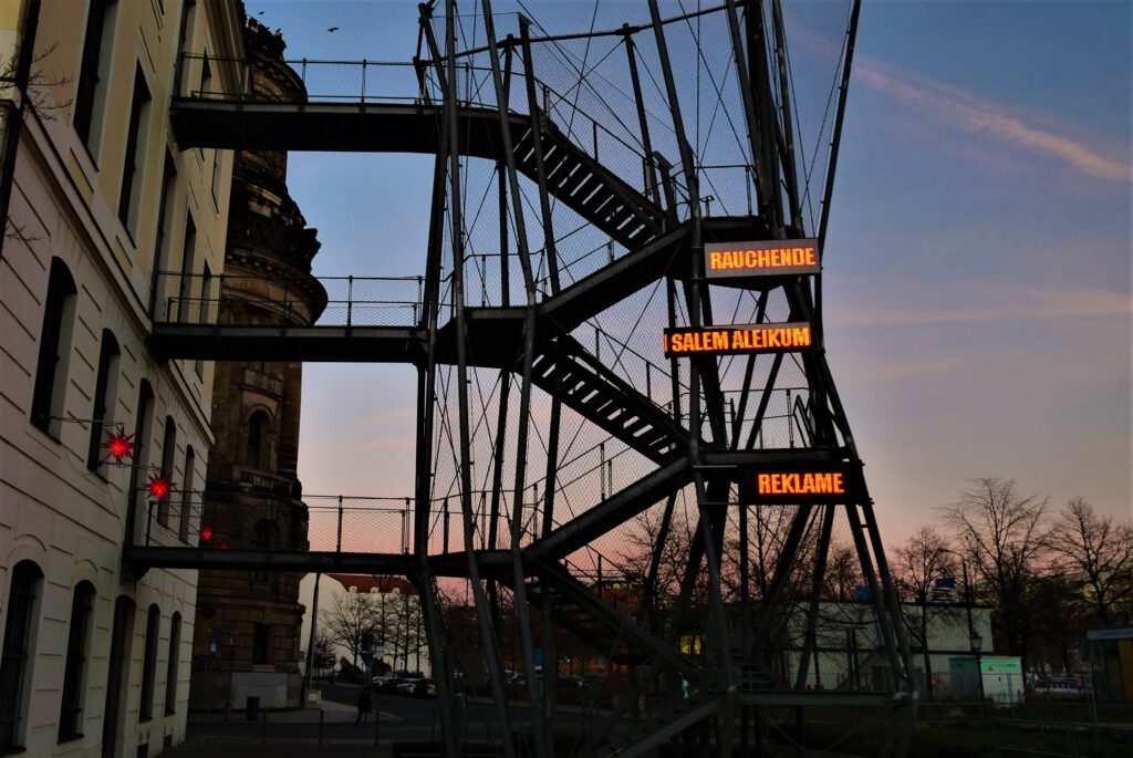 Eine LED-Installation an der Fluchttreppe des Landhauses zeigt aufgeteilt in drei Abschnitte untereinander die Worte "RAUCHENDE / SALEM ALEIKUM / REKLAME" 