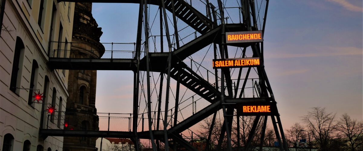 Eine Installation an der Fluchttreppe des Landhauses zeigt in drei LED-Panels untereinander die Worte "RAUCHENDE/ SALEM ALEIKUM / REKLAME". Die Worte leuchten rot und bewegen sich.