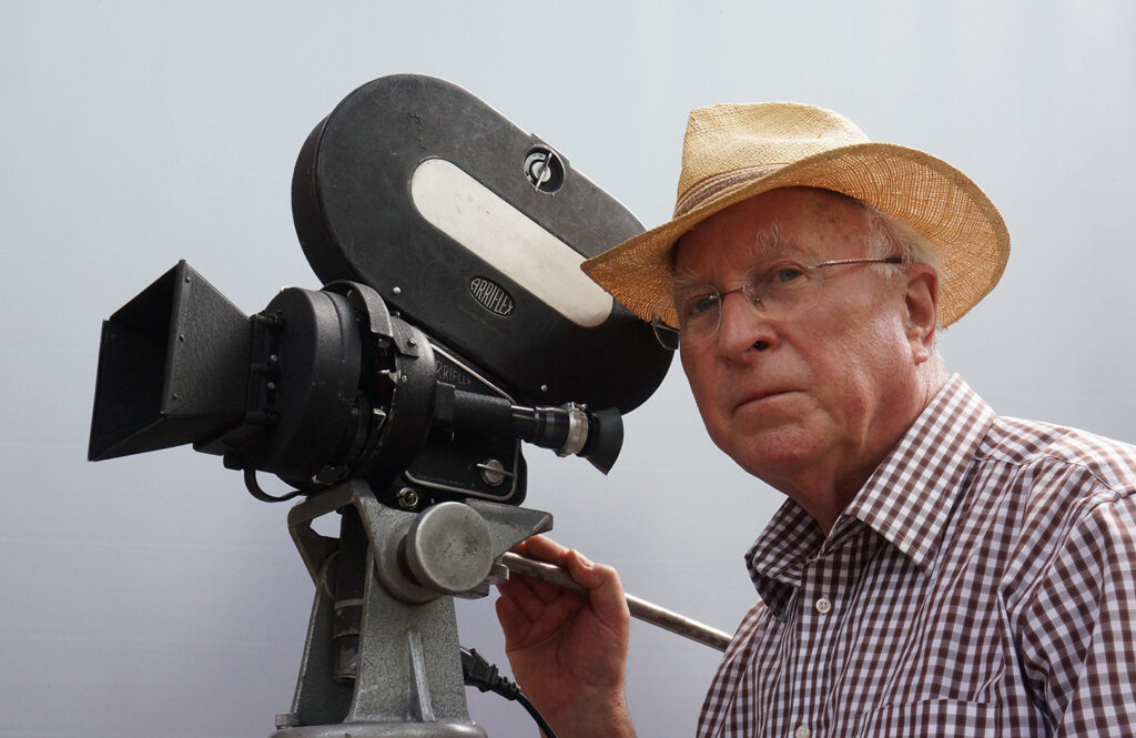 Farbaufnahme von Ernst Hirsch mit Strohut neben einer Kamera der Firma Arriflex.