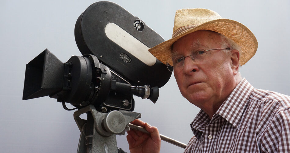 Farbaufnahme von Ernst Hirsch mit Strohut neben einer Kamera der Firma Arriflex.
