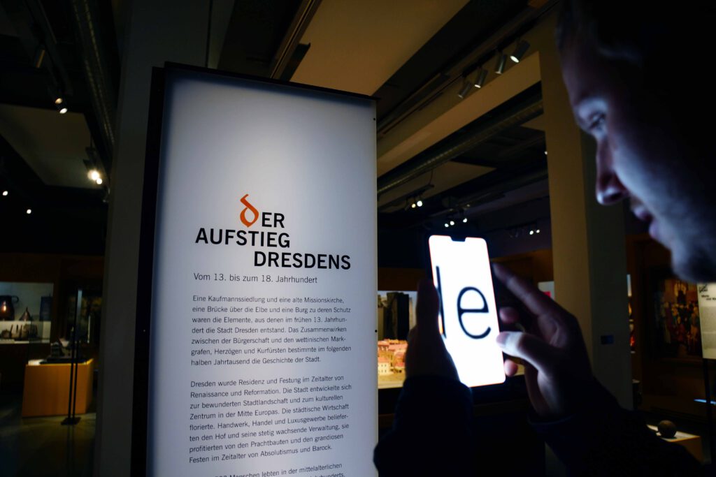 Ein Museumsbesucher liest einen Ausstellungstext mithilfe einer Kameraapp, die den Text auf dem Smartphone-Bildschirm stark vergrößert darstellt.