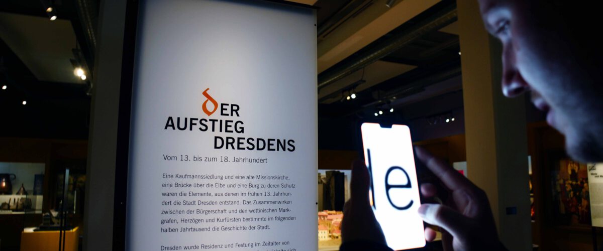 Ein Museumsbesucher liest einen Ausstellungstext mithilfe einer Kameraapp, die den Text auf dem Smartphone-Bildschirm stark vergrößert darstellt.