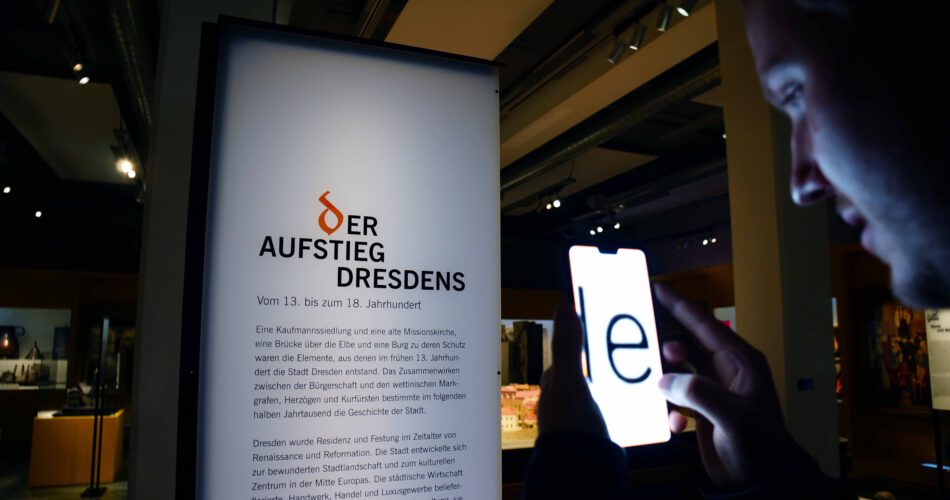 Ein Museumsbesucher liest einen Ausstellungstext mithilfe einer Kameraapp, die den Text auf dem Smartphone-Bildschirm stark vergrößert darstellt.