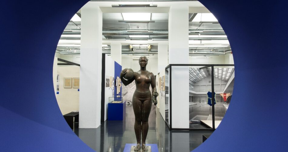 Eine Wand in kräftigem Blau. Durch eine große runde Aussparung schaut man direkt auf die dahinter stehende Wasserballspielerin in der Ausstellung.